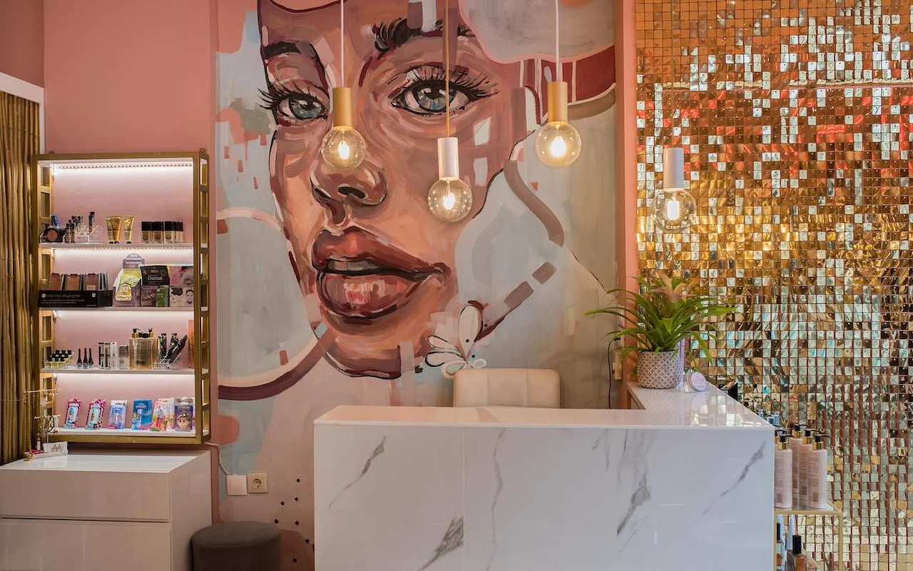 Een roze salon met een grote geschilderde muurschildering van een vrouwengezicht, een glinsterende gouden pailletmuur, een receptiebalie van wit marmer en hanglampen met blote peertjes.