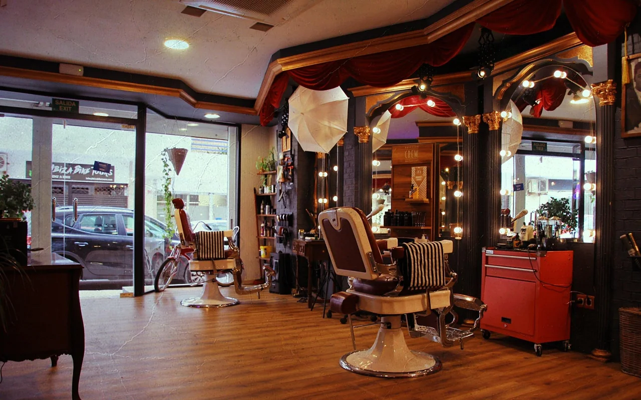 Een barbershop in vintage-stijl met klassieke leren ligstoelen op chromen voeten en gestreepte handdoeken. Vergulde spiegels met ronde peertjes-lampjes en rode fluwelen gordijnen staan langs de muur, een rood gereedschapskarretje staat opzij, en ramen aan de straatkant lichten de houten vloer op.