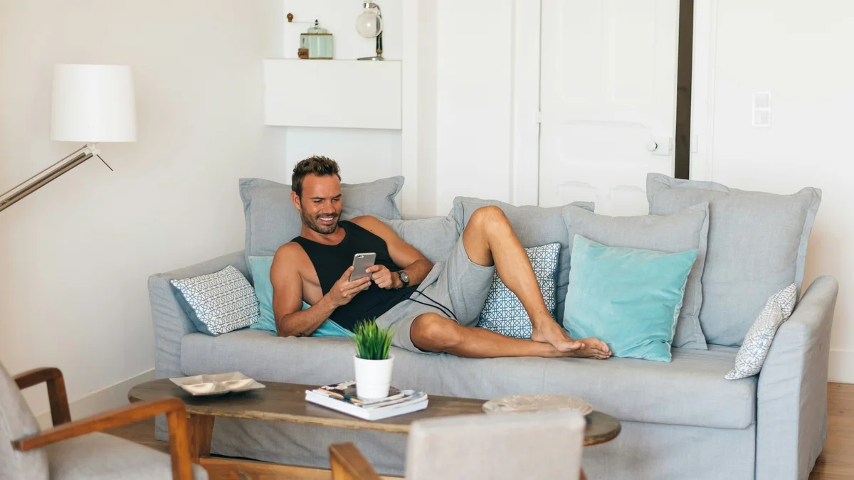 Een man ontspant met zijn voeten omhoog op zijn bank en lacht terwijl hij op zijn telefoon kijkt; hij draagt een zwart hemd en een losse, grijze short